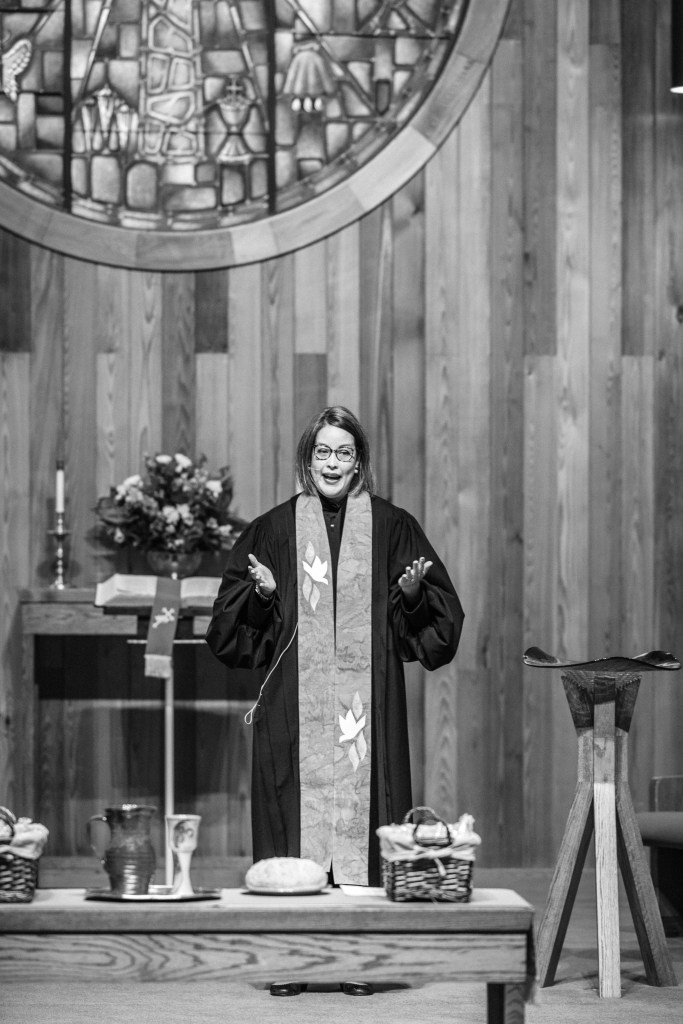 Black and white image of Crystal Varner Parker in clergy robes and stole in a church sanctuary with a communion table in front of her and baptismal font beside her.